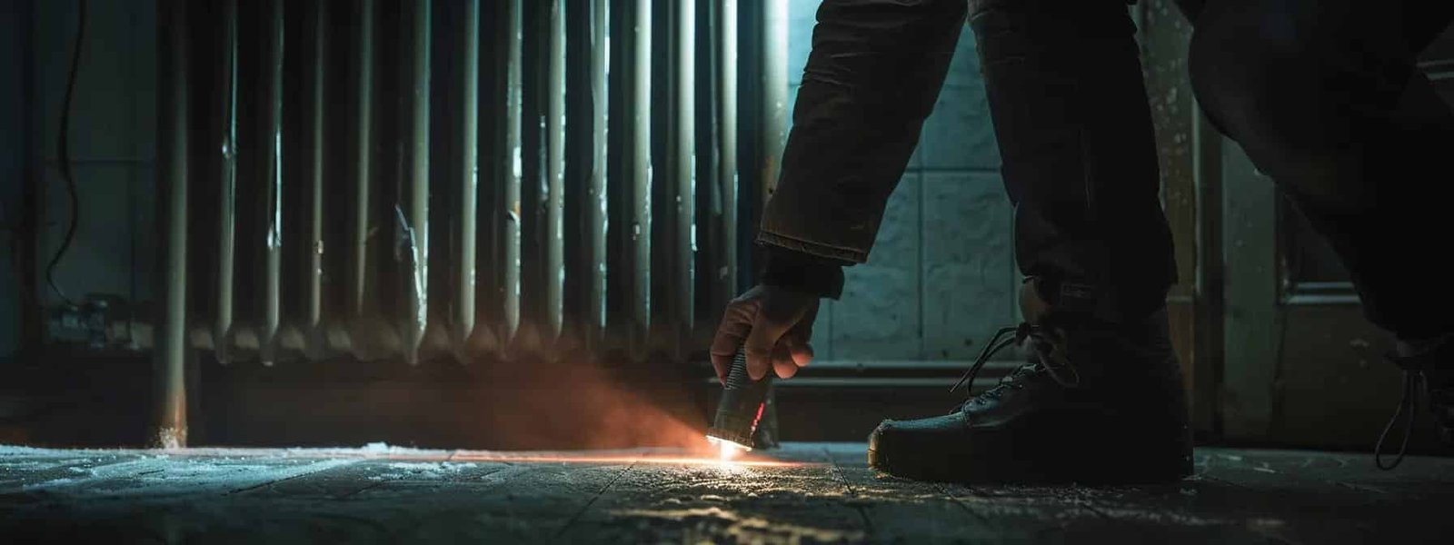 a hand holding a flashlight, kneeling in front of a dark heater in a dimly-lit room, inspecting its power source.